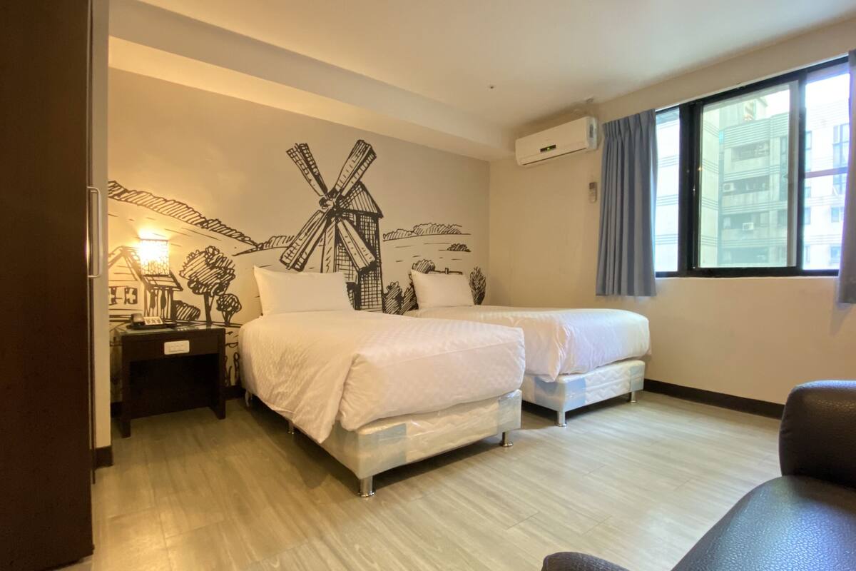 A hotel room with two beds, a desk with a lamp, a large window with curtains, and a windmill mural on the wall.