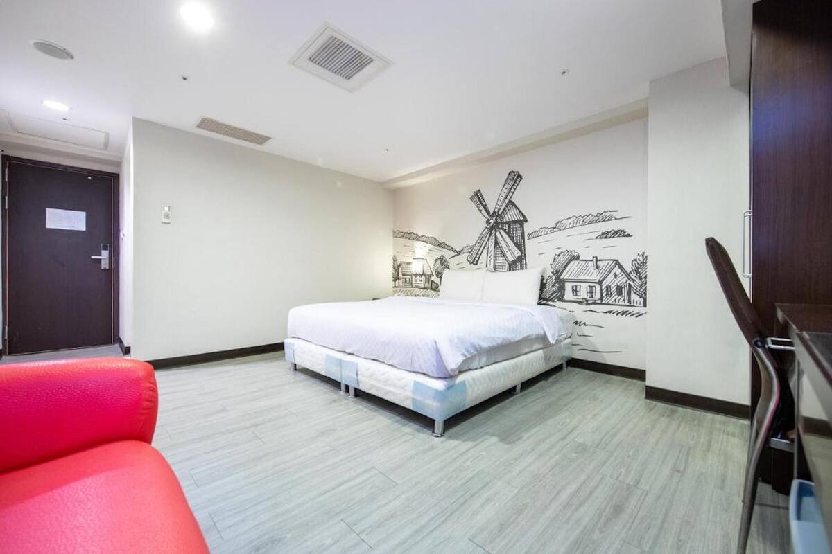 A hotel room with a bed, a red chair, a wooden desk, and a mural of windmills on the wall.