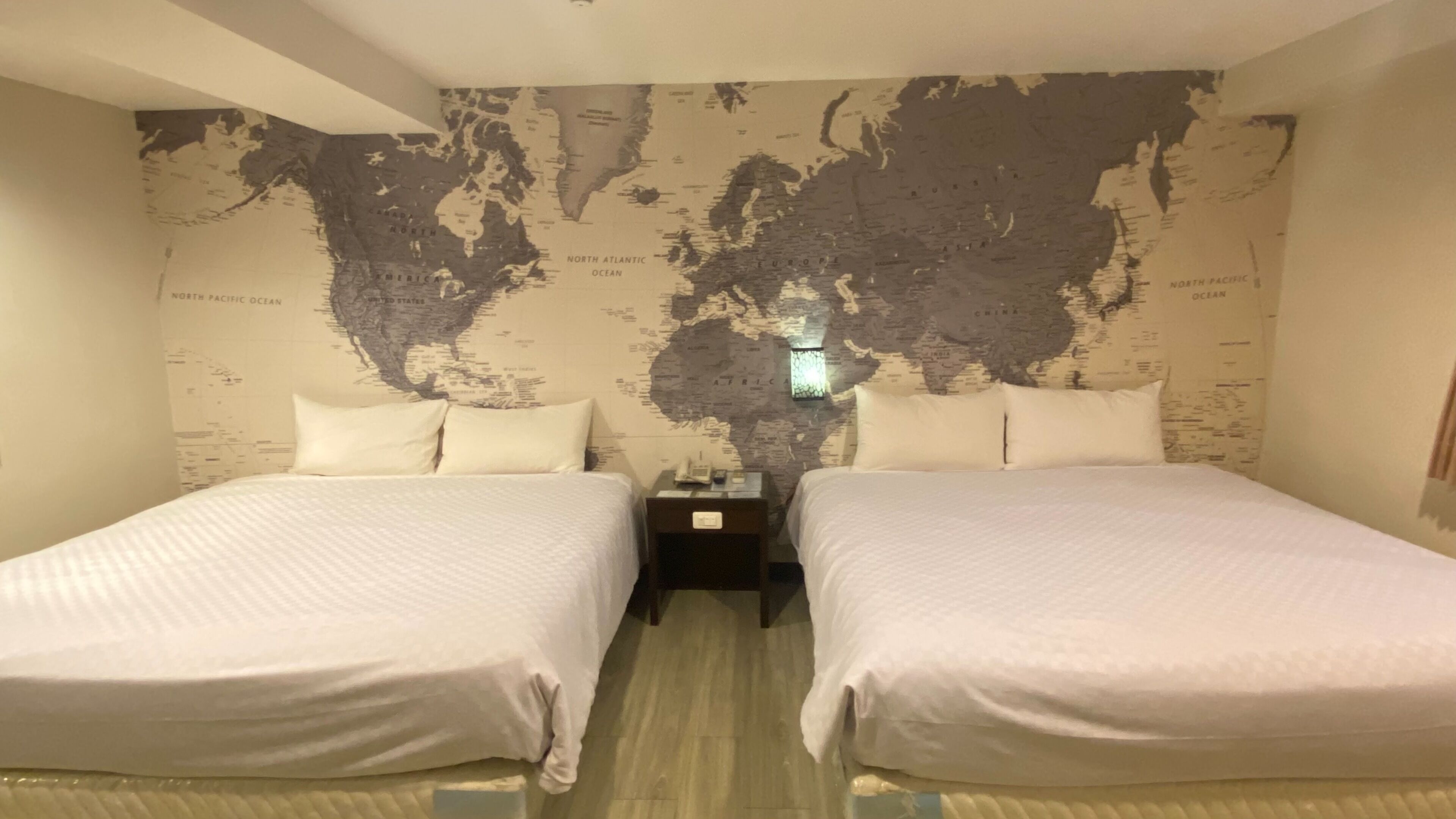 A hotel room with two beds, a world map on the wall, and a nightstand with a phone.
