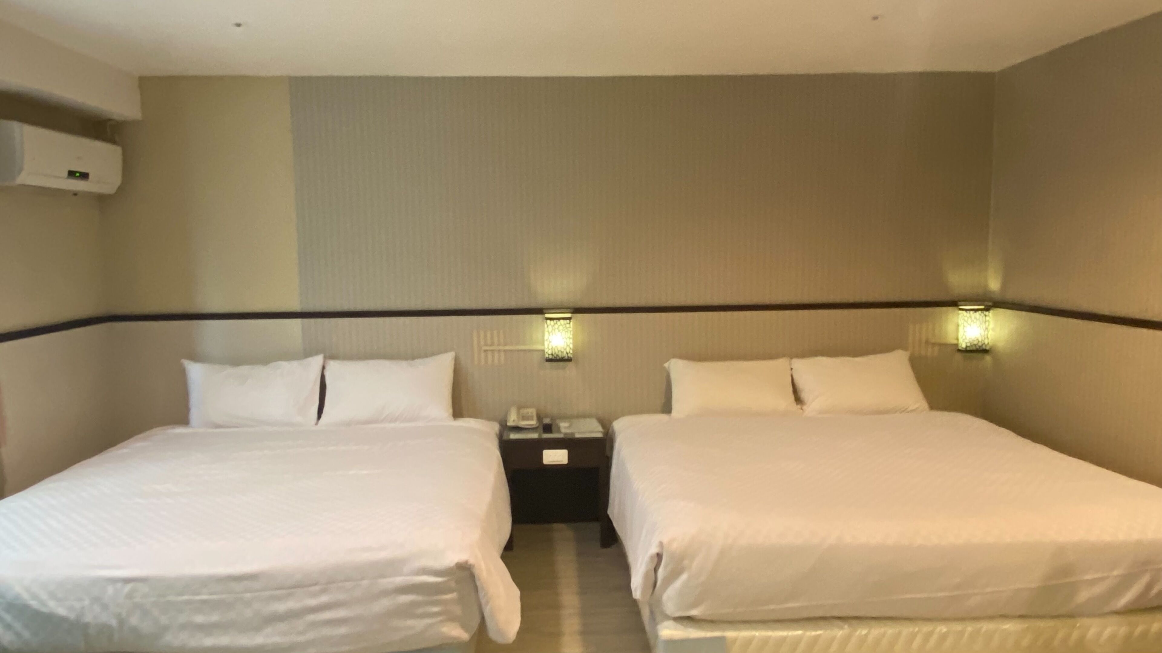 Een hotelkamer met twee bedden, een nachtkastje met een telefoon en wandlampen.