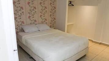 A small, neatly made bed with a white mattress and pillows, a floral wallpaper pattern on the wall behind it, and a simple, uncluttered room layout.
