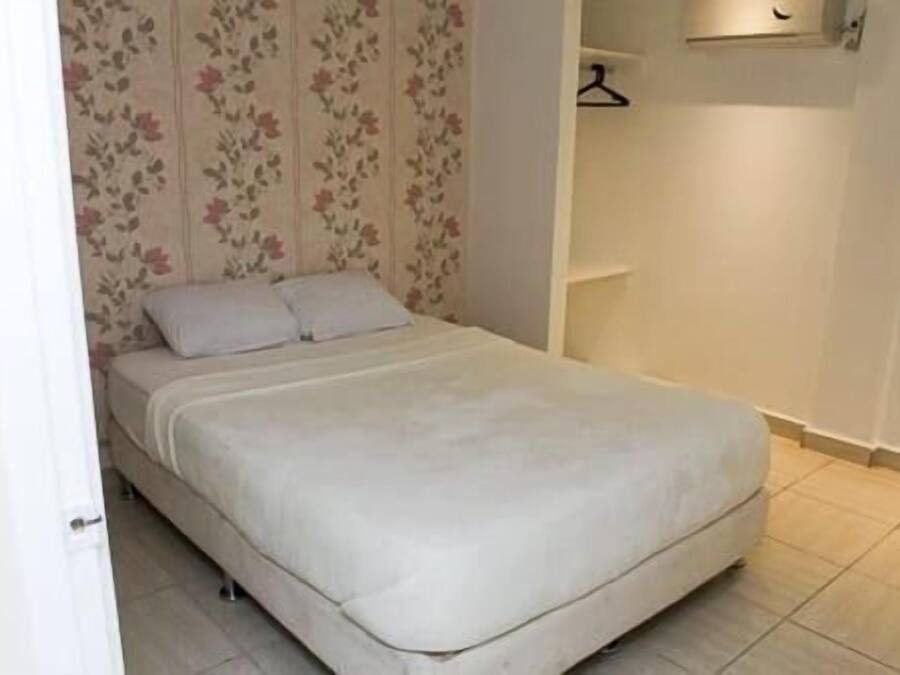 A small, neatly made bed with a white mattress and pillows, a floral wallpaper pattern on the wall behind it, and a simple, uncluttered room layout.