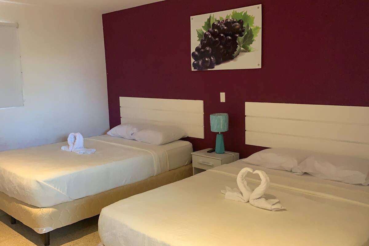 Two single beds with white linens, a teal nightstand with a lamp, and a wall-mounted picture of grapes.
