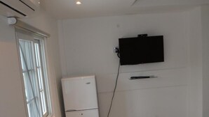 Una habitación pequeña y limpia con aire acondicionado blanco, refrigerador blanco, televisor de pantalla plana montado en la pared y una cama con colchón blanco.