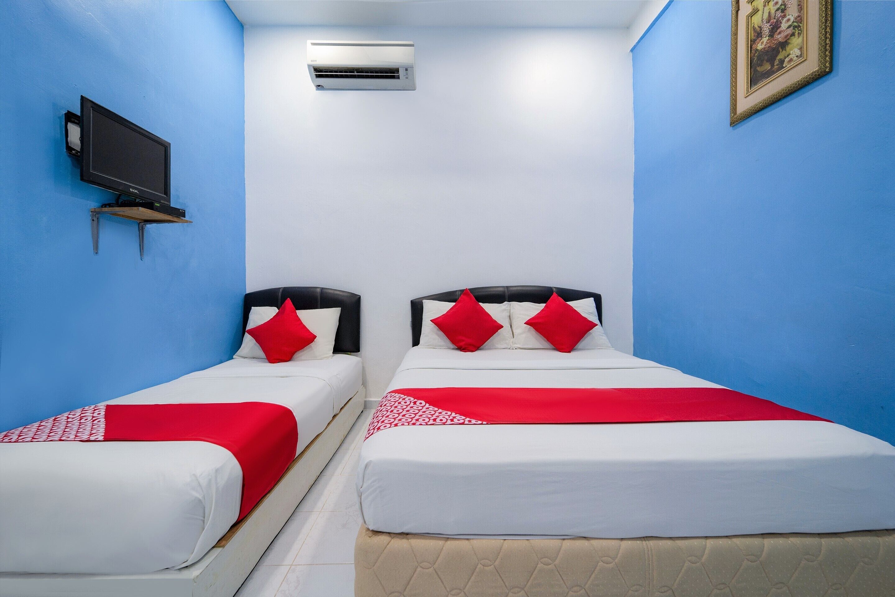 A hotel room with two beds, each with red and white bedding, a flat-screen TV on a stand, and an air conditioning unit on the wall.