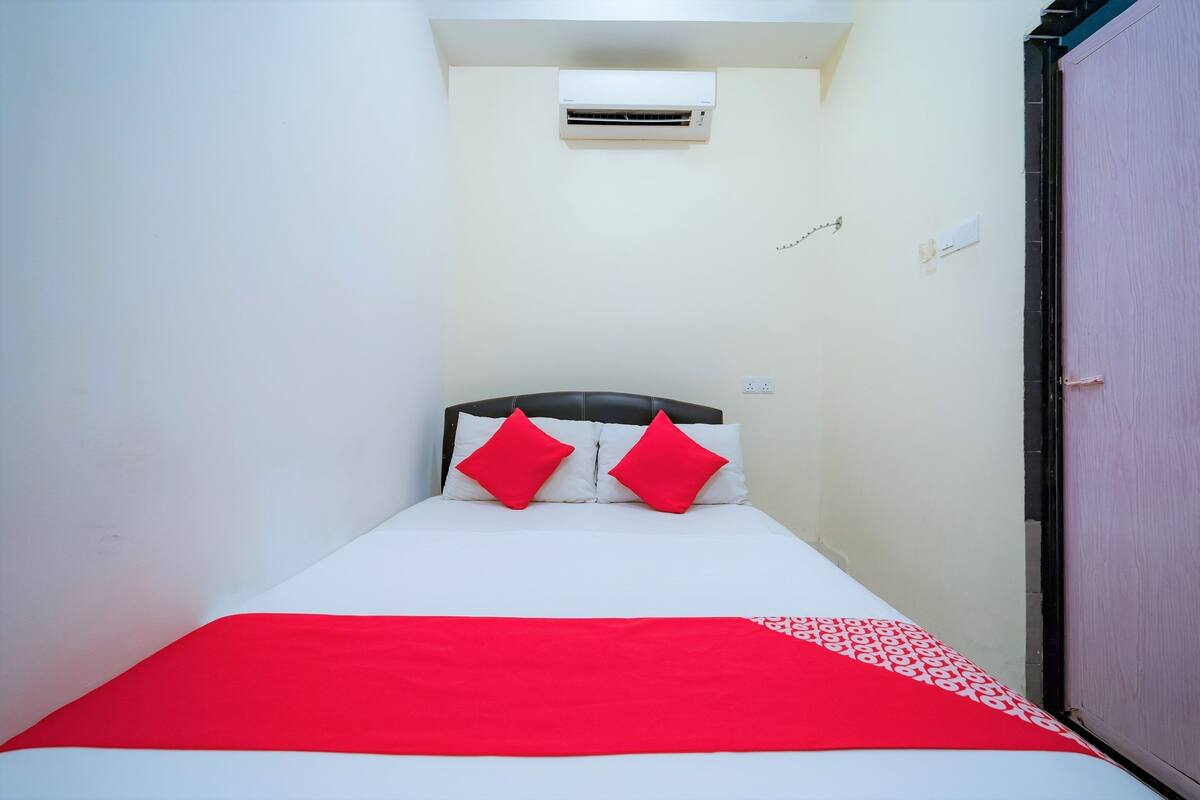 A small, narrow hotel room with a single bed, white and red bedding, and a red and white patterned throw.