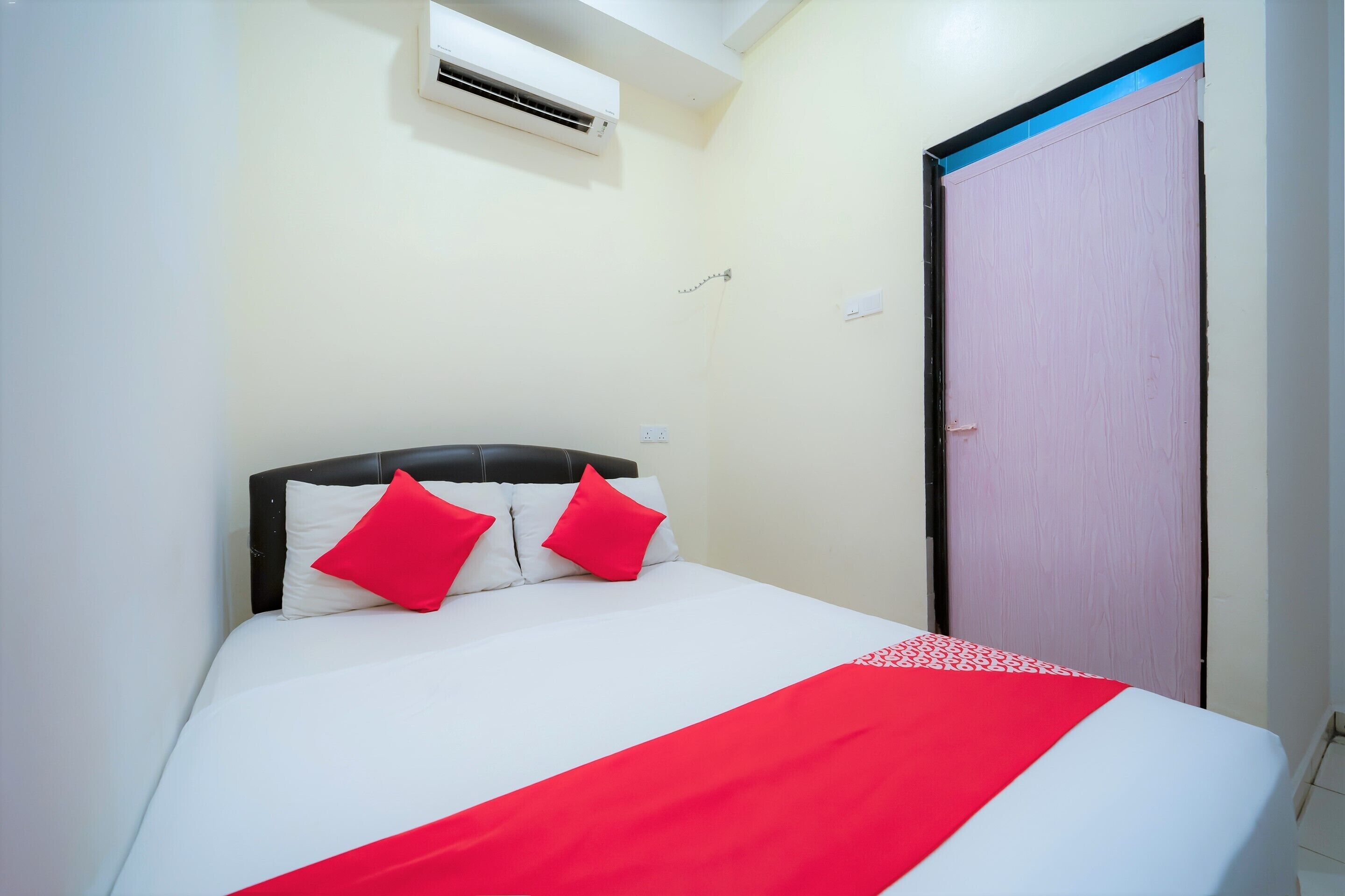 A bed with white bedding and red pillows, an air conditioning unit above it, and a door to the right.