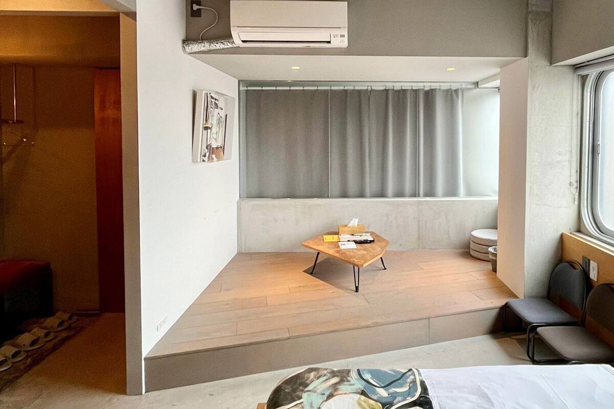 A modern hotel room with a bed, a small table, a window with curtains, an air conditioning unit, and a bathroom with a toilet and sink.