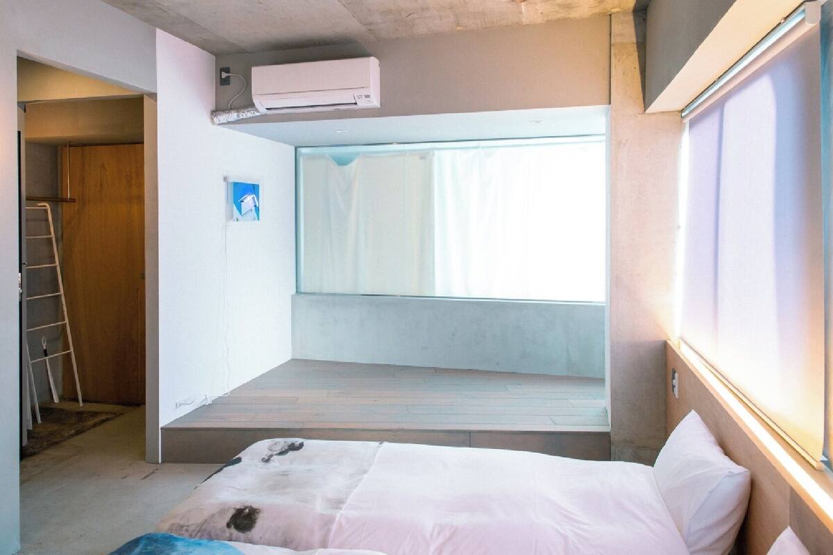 A compact room with a bed, a window with curtains, an air conditioning unit, and a ladder against the wall.