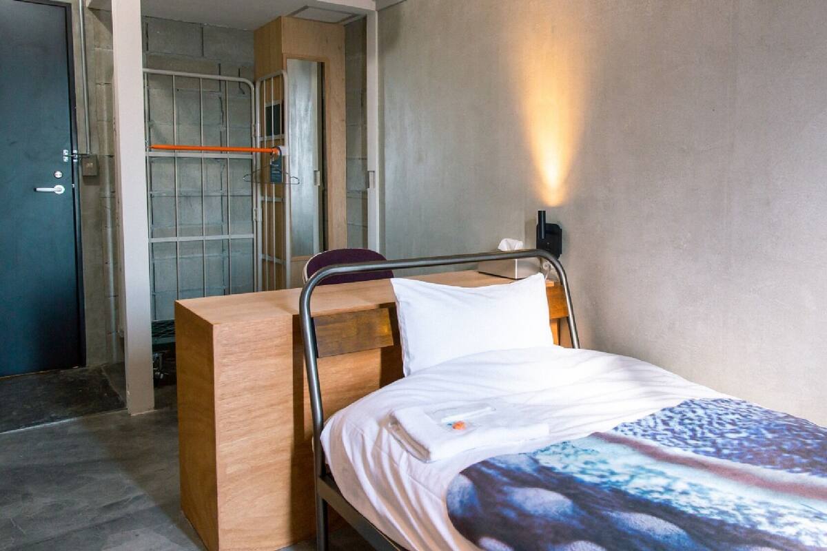 A single bed with a metal frame, a wooden bedside table, and a wall-mounted light fixture.