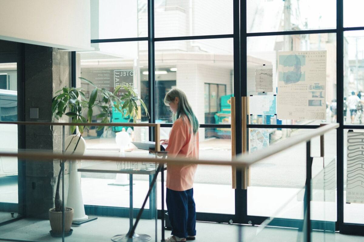 A person standing near a window with a view of a cityscape, a potted plant, and a table with a laptop.