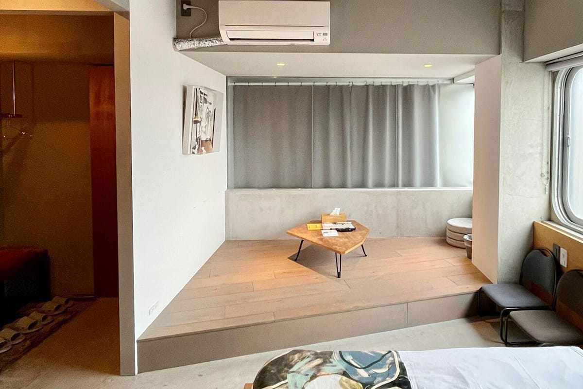 Habitación de hotel pequeña con cama, una mesita, ventana con cortinas, unidad de aire acondicionado y una puerta que da a otra habitación.