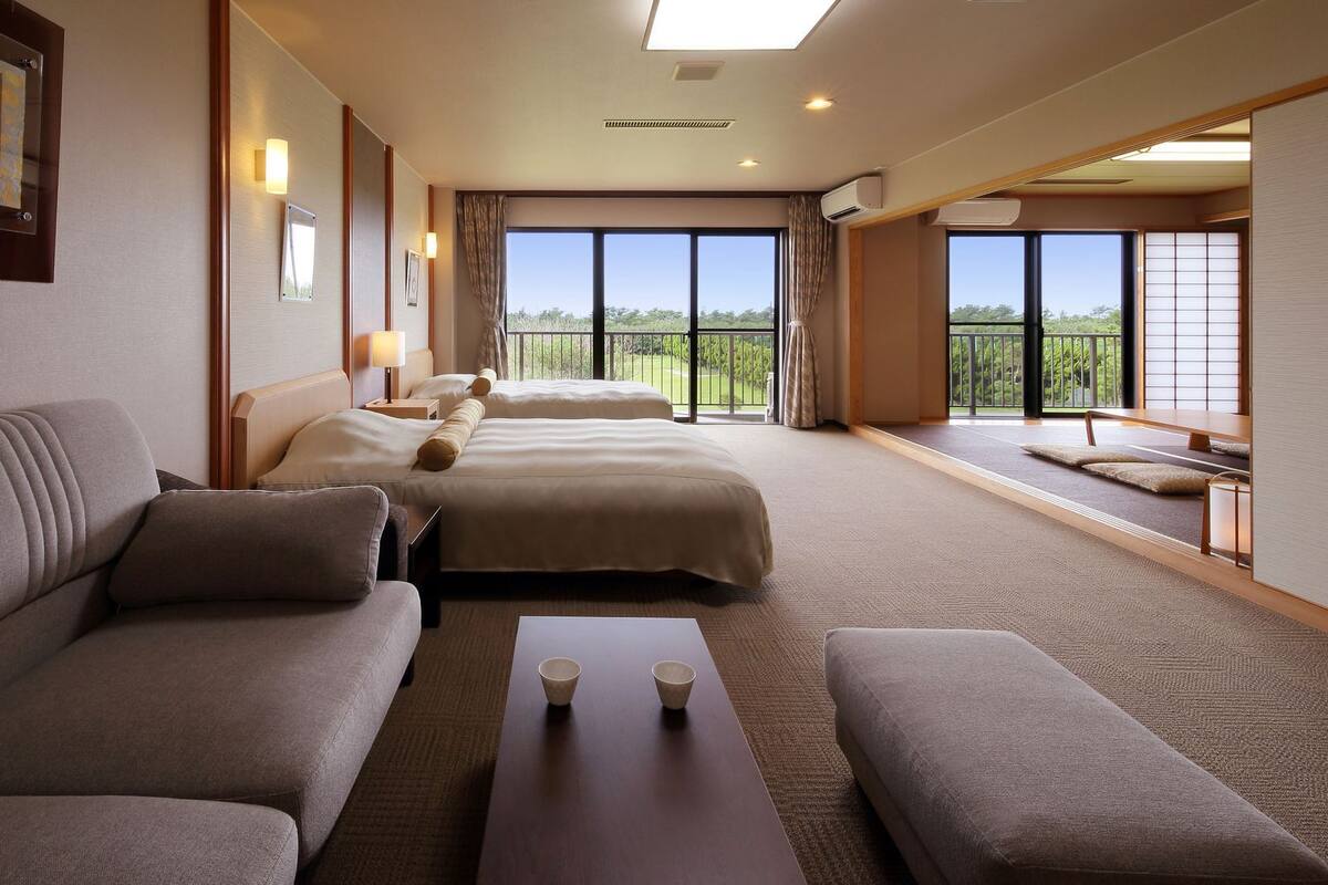 A hotel room with a sofa, two beds, a small table, and a view of the outdoors.