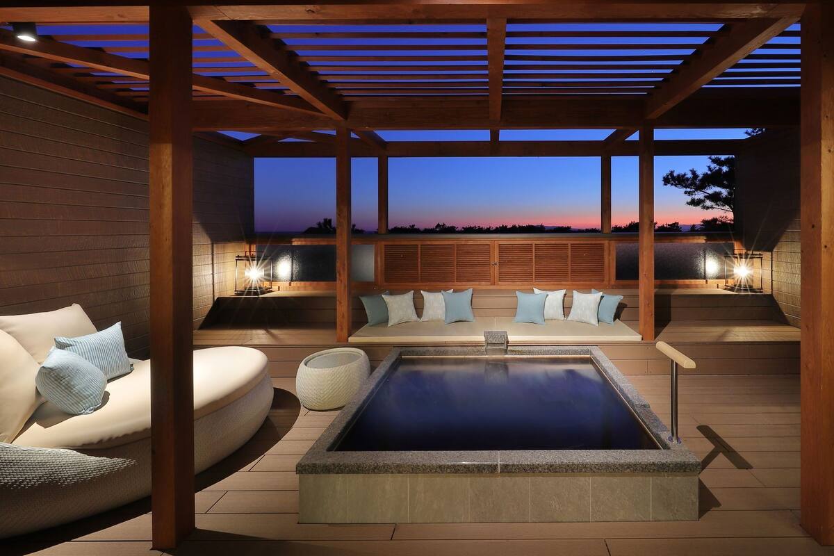 A covered outdoor area with a hot tub, seating, and a view of the sunset.
