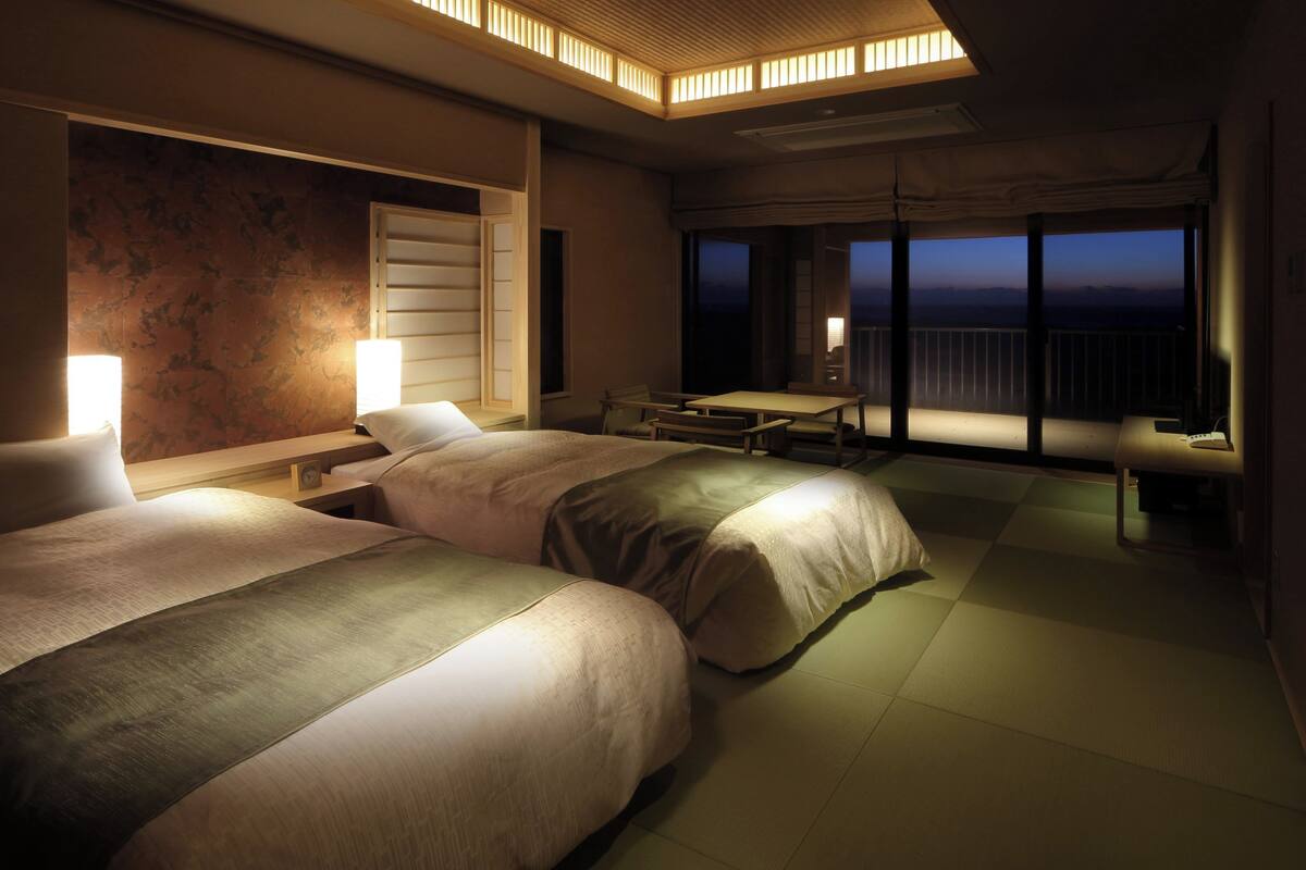 A hotel room with two beds, a desk, a chair, and a balcony with a view.