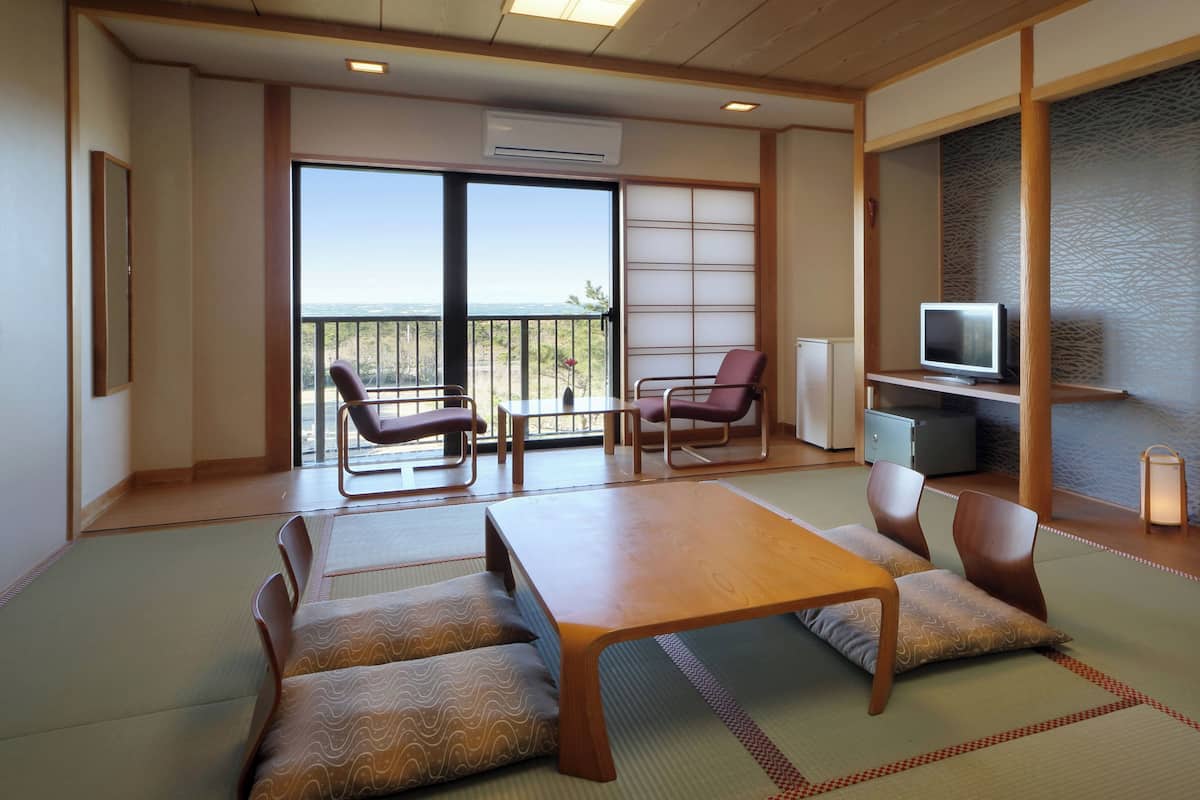 A traditional Japanese room with tatami flooring, a low table, cushioned seating, a television, and a view through a sliding door.