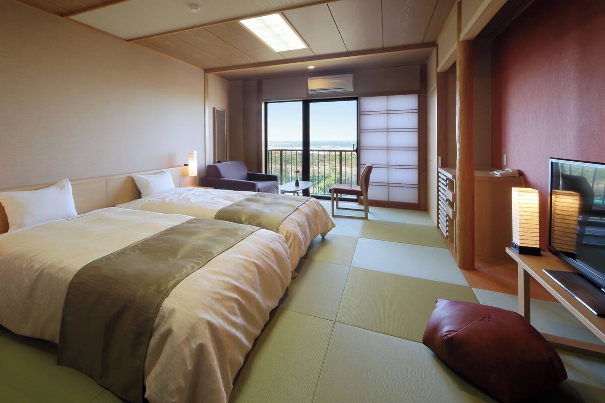 A hotel room with two beds, a TV, a small table, and a balcony with a view.