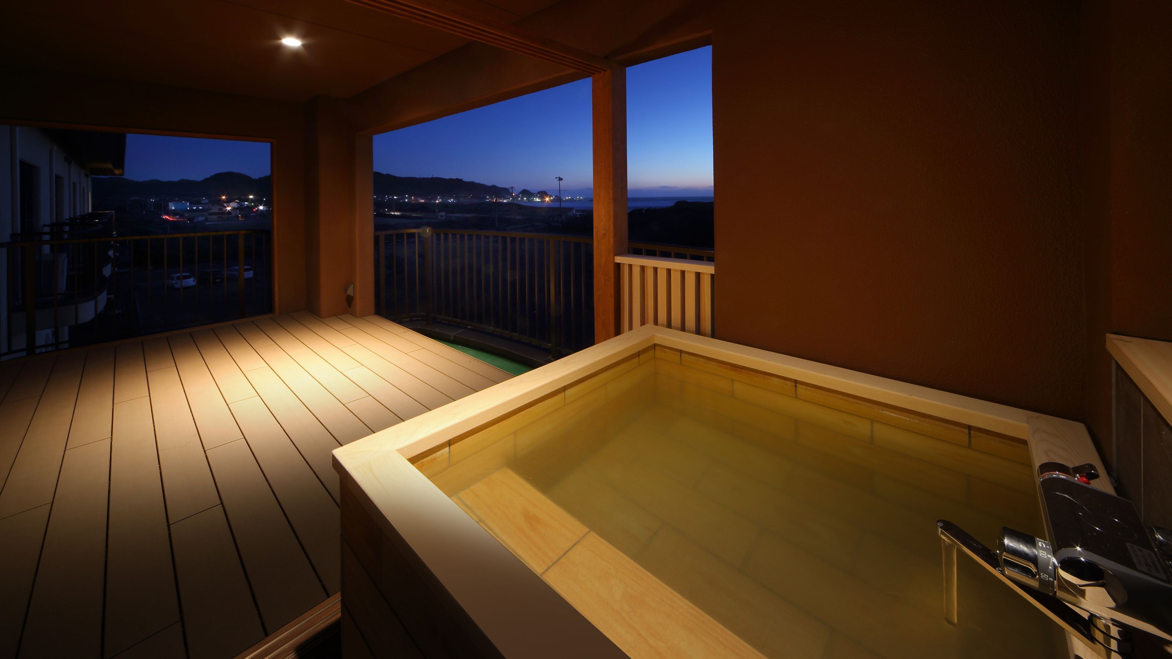 A wooden deck with a hot tub, a balcony with a view of the city at dusk, and a railing.