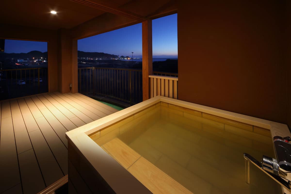 A wooden deck with a hot tub, a balcony with a view of the city at dusk, and a railing.