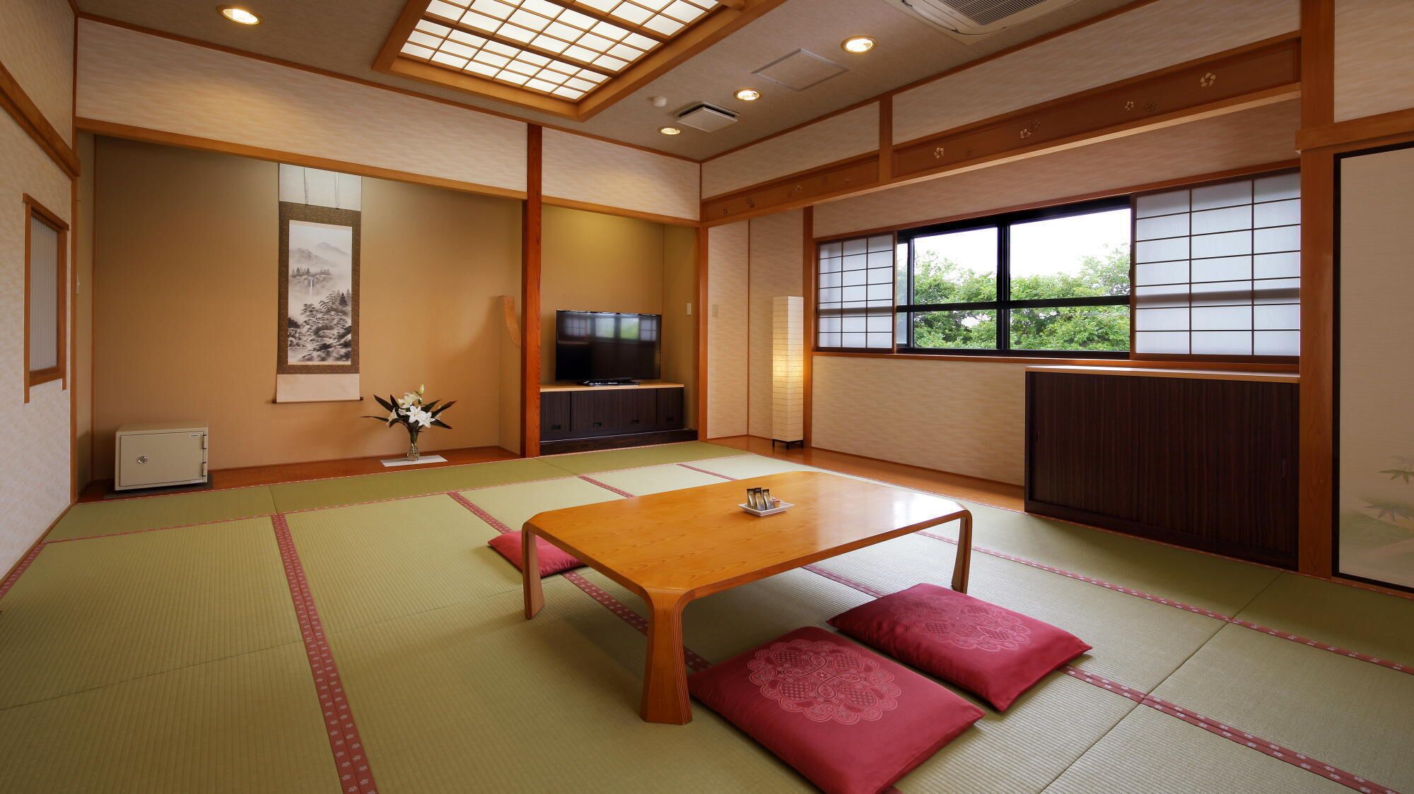 A traditional Japanese room with tatami flooring, a low wooden table, red cushions, a television, and a window with a view of greenery.