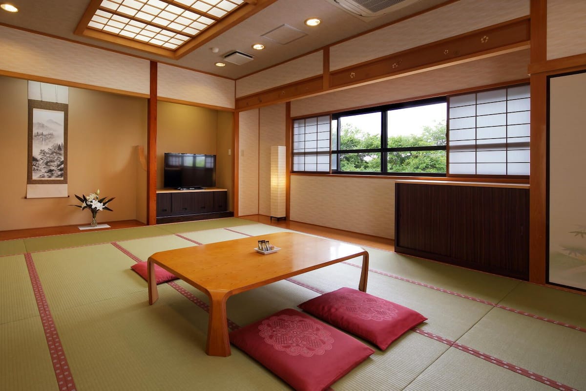 A traditional Japanese room with tatami flooring, a low wooden table, red cushions, a television, and a window with a view of greenery.