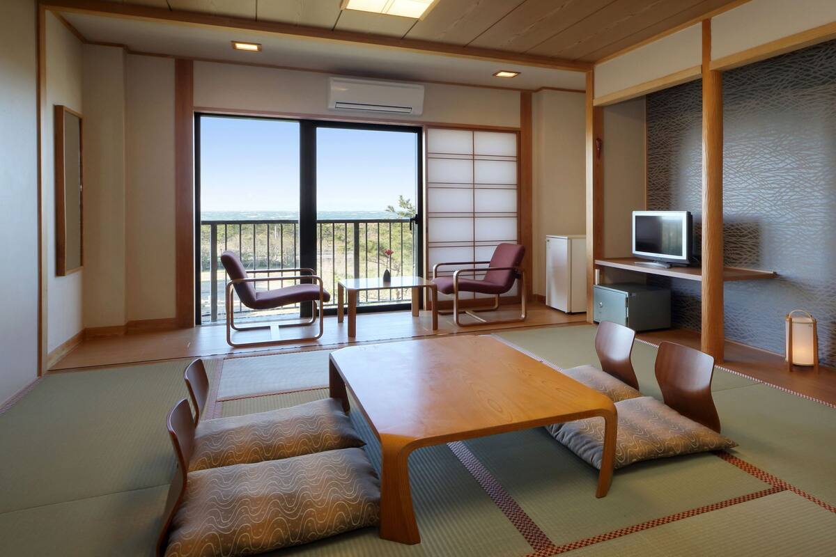 A traditional Japanese-style room with tatami flooring, a low table, cushioned seating, a television, and a view through a sliding door.