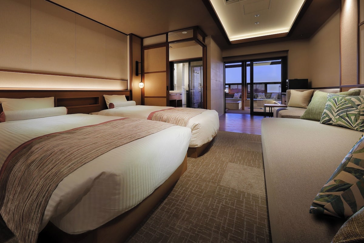 A hotel room with two beds, a sofa, a desk, and a view of the outside.