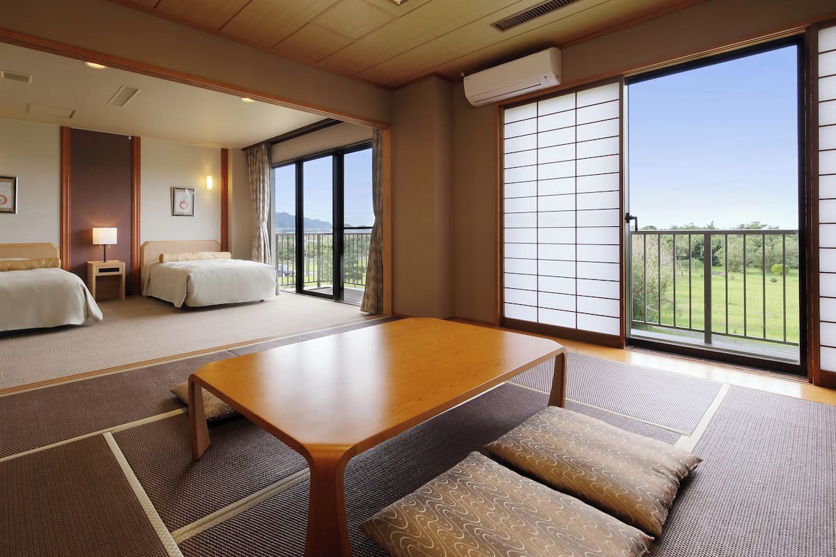 A hotel room with two beds, a wooden table, a sofa, a balcony with a view, and an air conditioning unit.