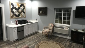 A modern kitchen with white cabinets, a microwave, and a sink. There's a decorative rug, a chair, and a television mounted on the wall.