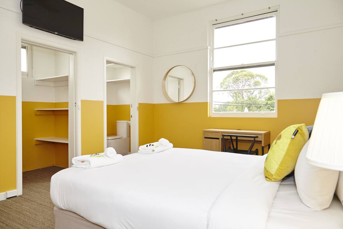 A hotel room with a bed, a desk, a chair, a mirror, and a window with a view of trees.