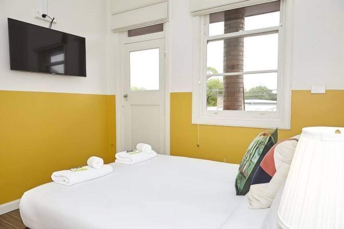 A hotel room with a bed, two windows, a flat-screen TV, and a view of greenery outside.