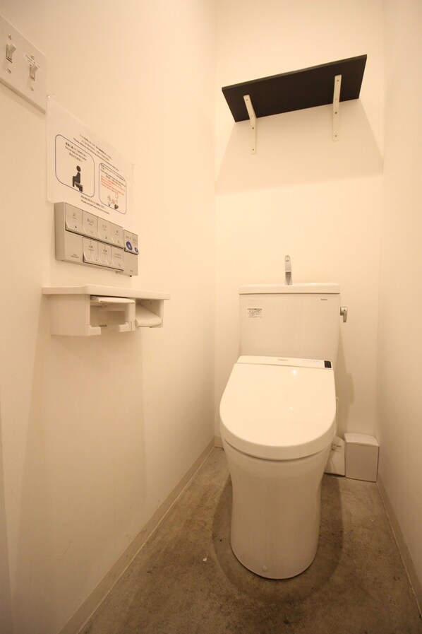 A small, clean bathroom with a toilet, a shelf with a sign, and a flush handle. - The Next Door - Hostel (Kyoto)