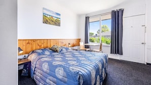 Un dormitorio con una cama grande, un cabecero de madera, una ventana con cortinas, un escritorio con lámpara y un cuadro en la pared.