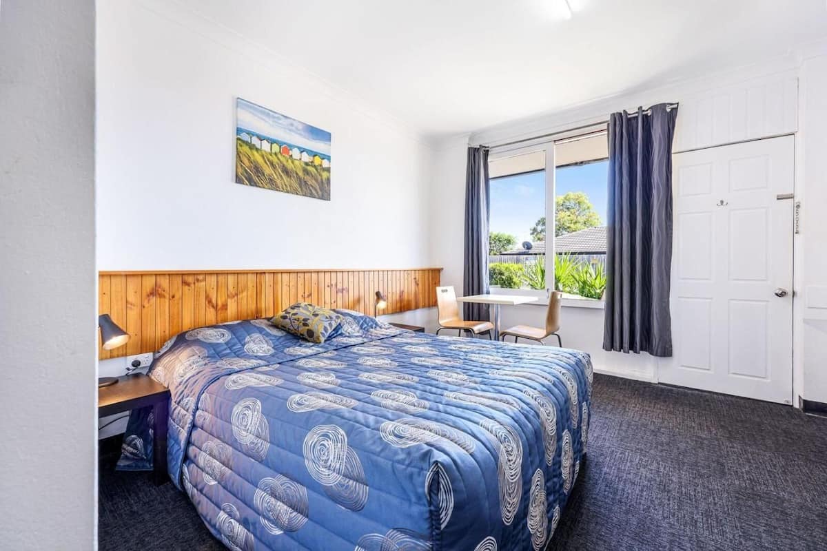 A bedroom with a large bed, a wooden headboard, a window with curtains, a desk with a lamp, and a painting on the wall.