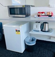 Una kitchenette compacta con microondas, un refrigerador pequeño, un tostador y una tetera sobre una encimera blanca.