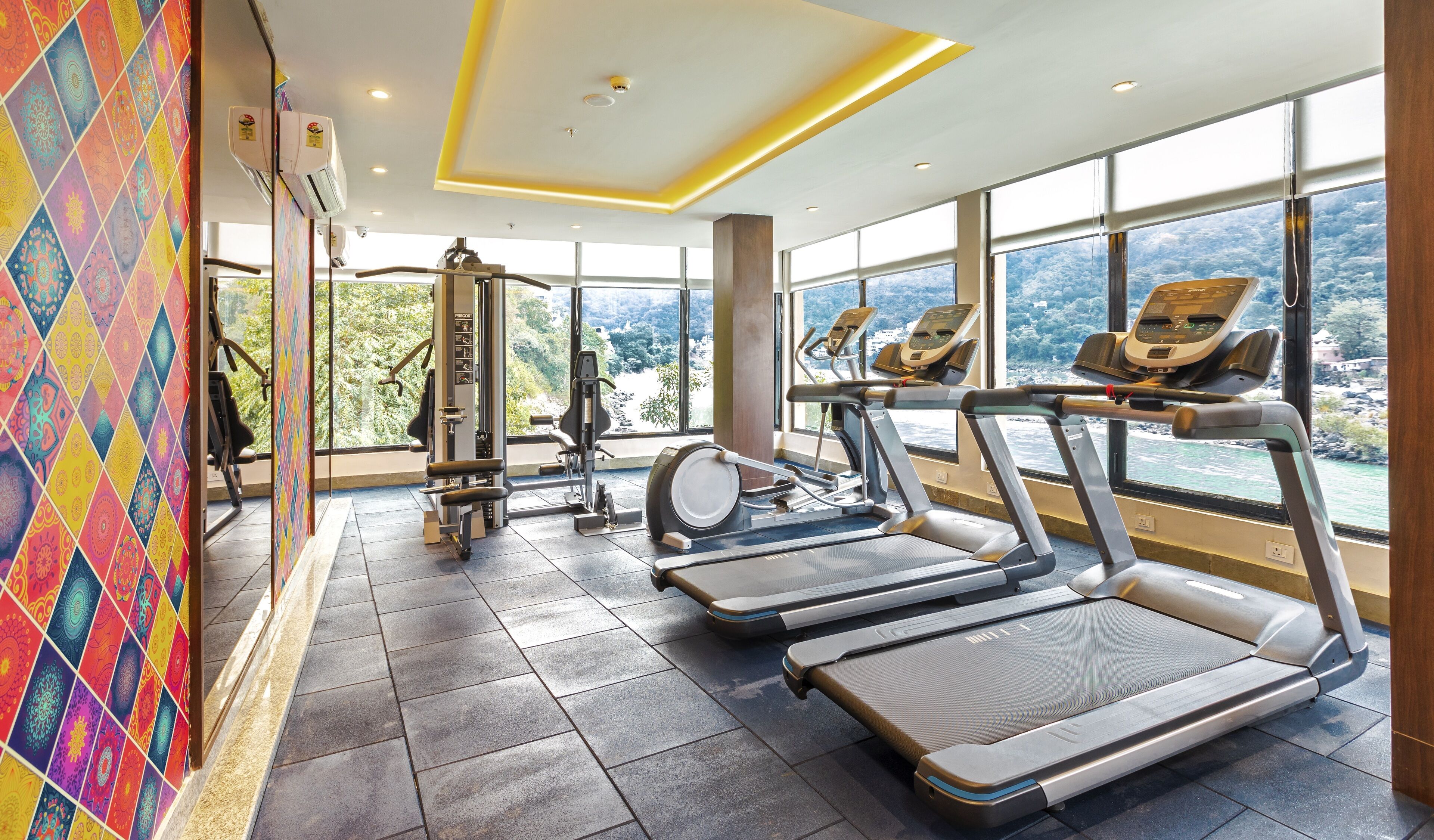 Un gimnasio con cintas de correr, una máquina de remo y un banco de pesas. Amplias ventanas ofrecen vistas a un paisaje montañoso.