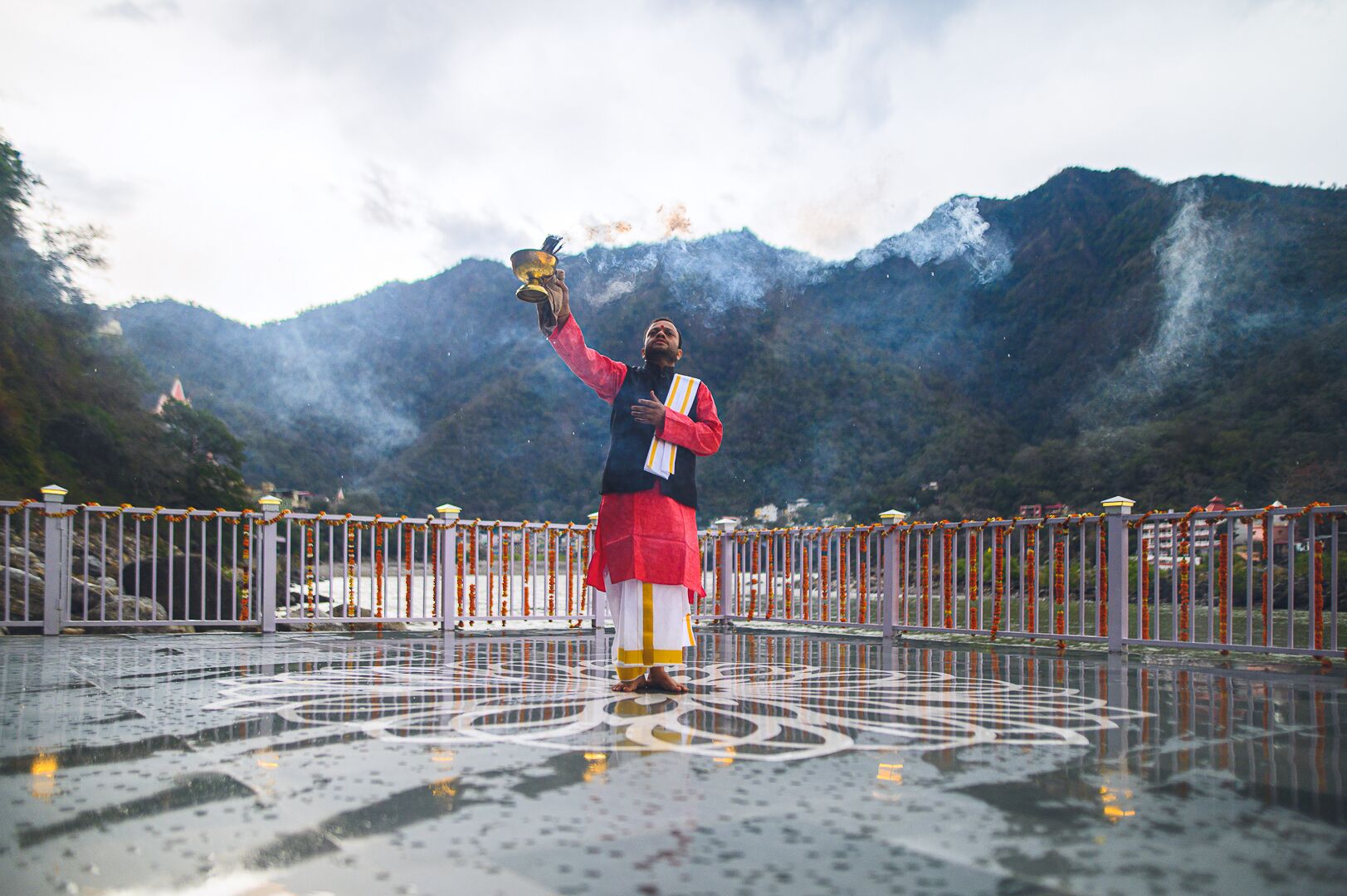 A person standing on a bridge with a mountainous backdrop, holding a ceremonial object.