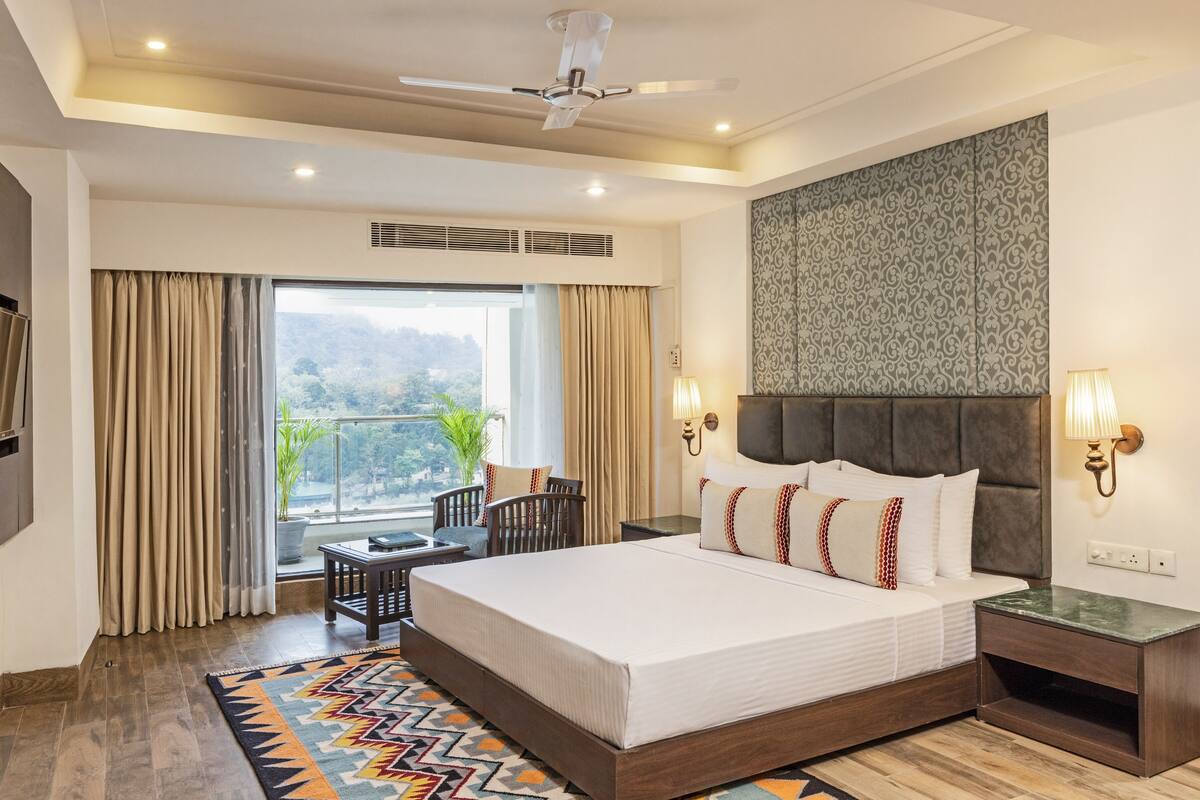 A hotel room with a large bed, a ceiling fan, a view of the outdoors, a small table with chairs, and a nightstand with a lamp.