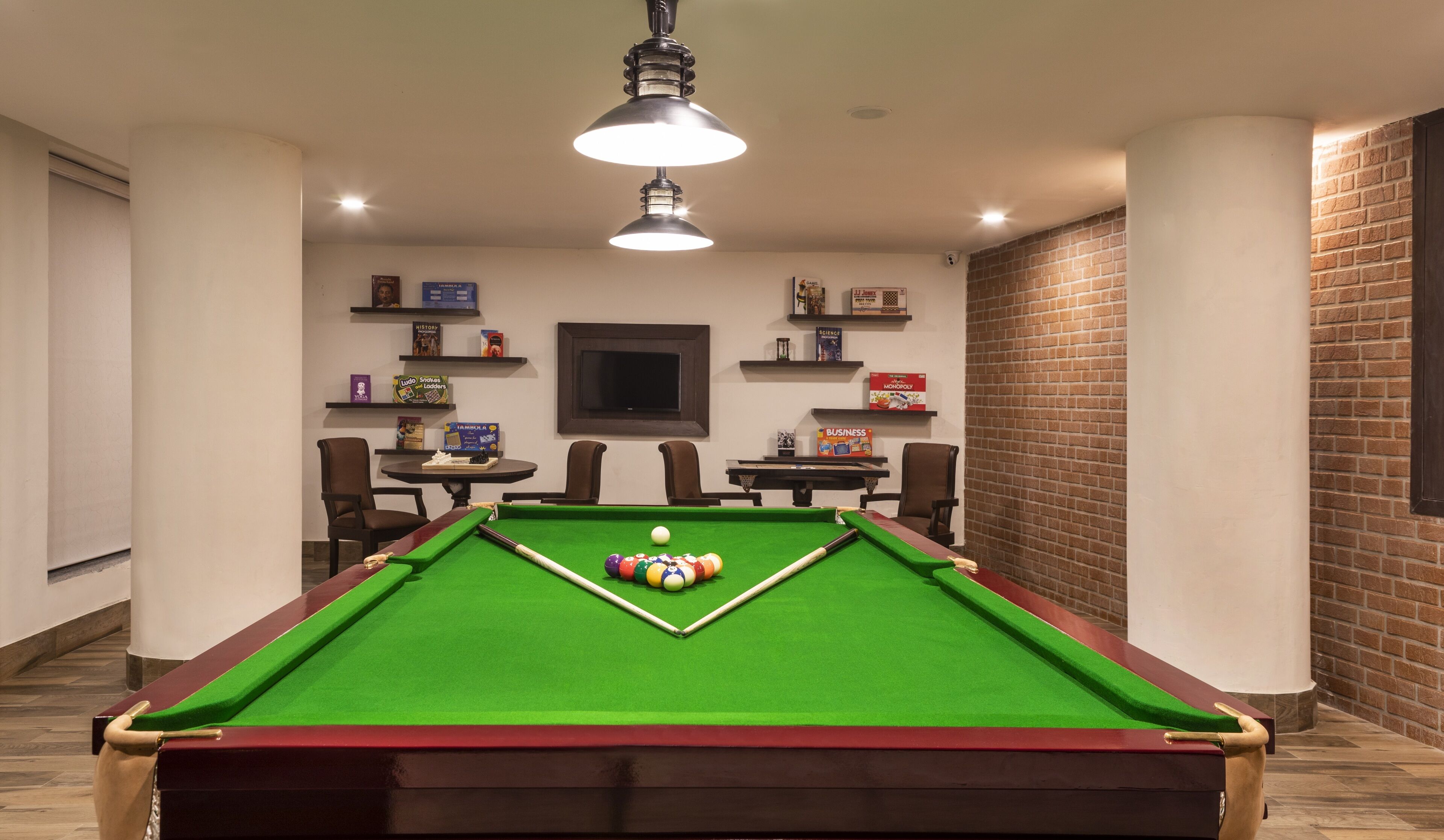Una mesa de billar con superficie de fieltro verde, rodeada por cuatro sillas, un televisor montado en la pared y estantes con varios juegos de mesa y libros.