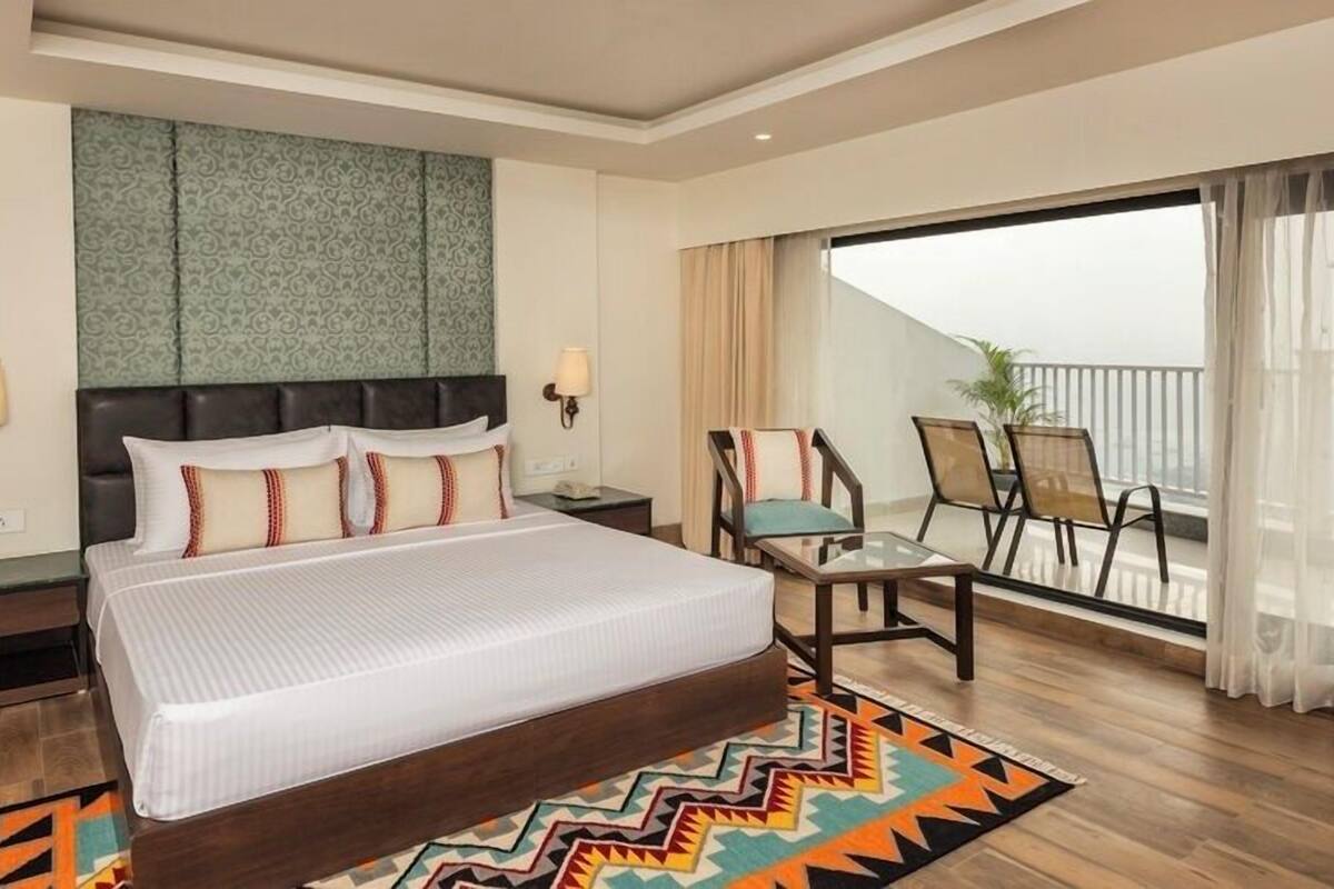 A hotel room with a large bed, a small table, two chairs, a balcony with a view, and a patterned wall.