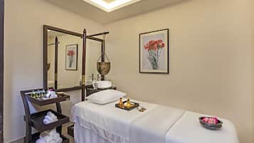 A spa room with a massage table, a wooden side table with towels and toiletries, a large mirror, and a framed floral artwork on the wall.