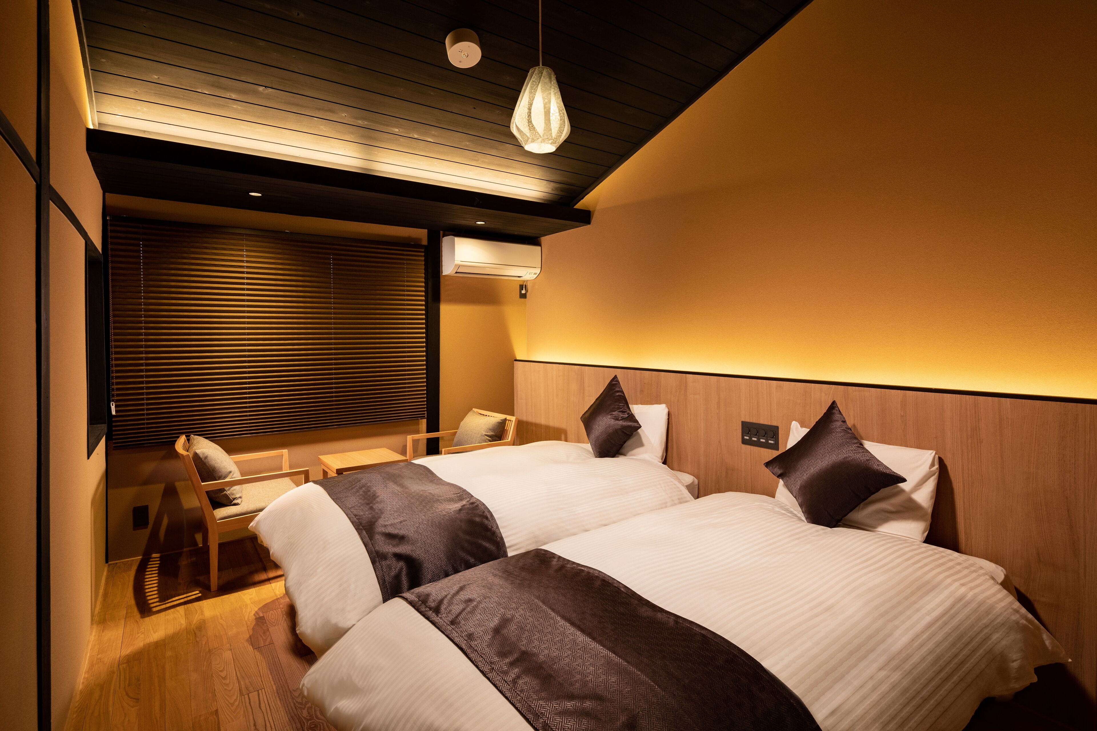 A hotel room with two beds, a wooden headboard, a bedside table, a chair, and a window with blinds.
