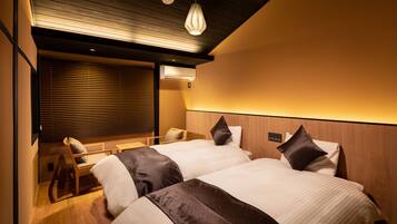 A hotel room with two beds, a wooden headboard, a bedside table, a chair, and a window with blinds.