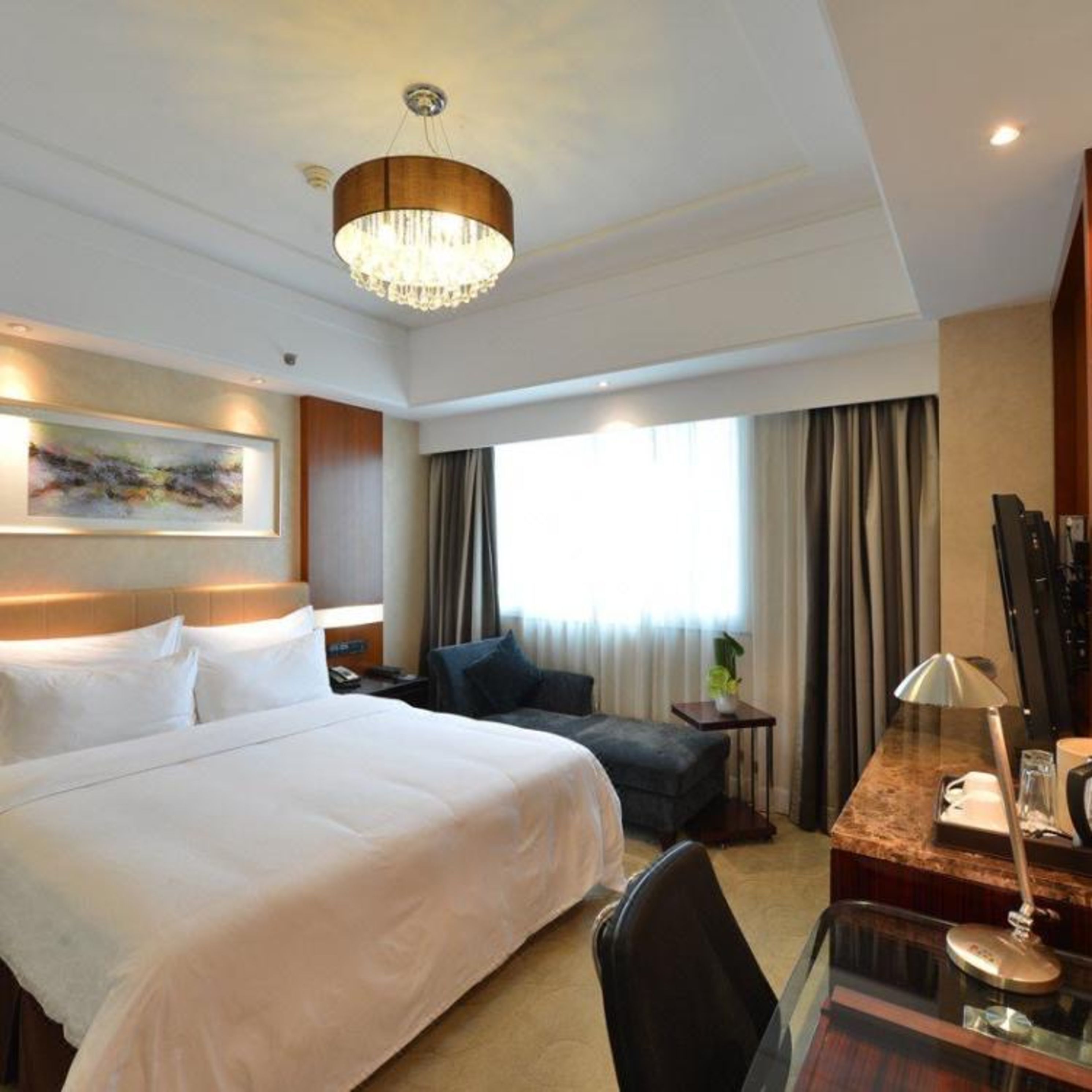 A hotel room with a large bed, a desk with a lamp, a chair, a small table with a vase, and a painting on the wall.