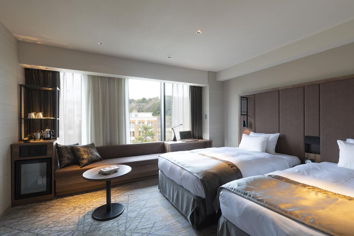 A hotel room with two beds, a sofa, a small table, a TV, and a window with curtains.