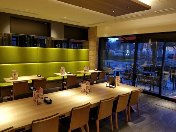 A modern dining area with a long wooden table, cushioned seating, and a view of the outdoors.