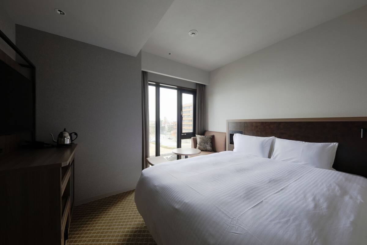 A hotel room with a large bed, a small table, a chair, a kettle, and a window with a view of the city.