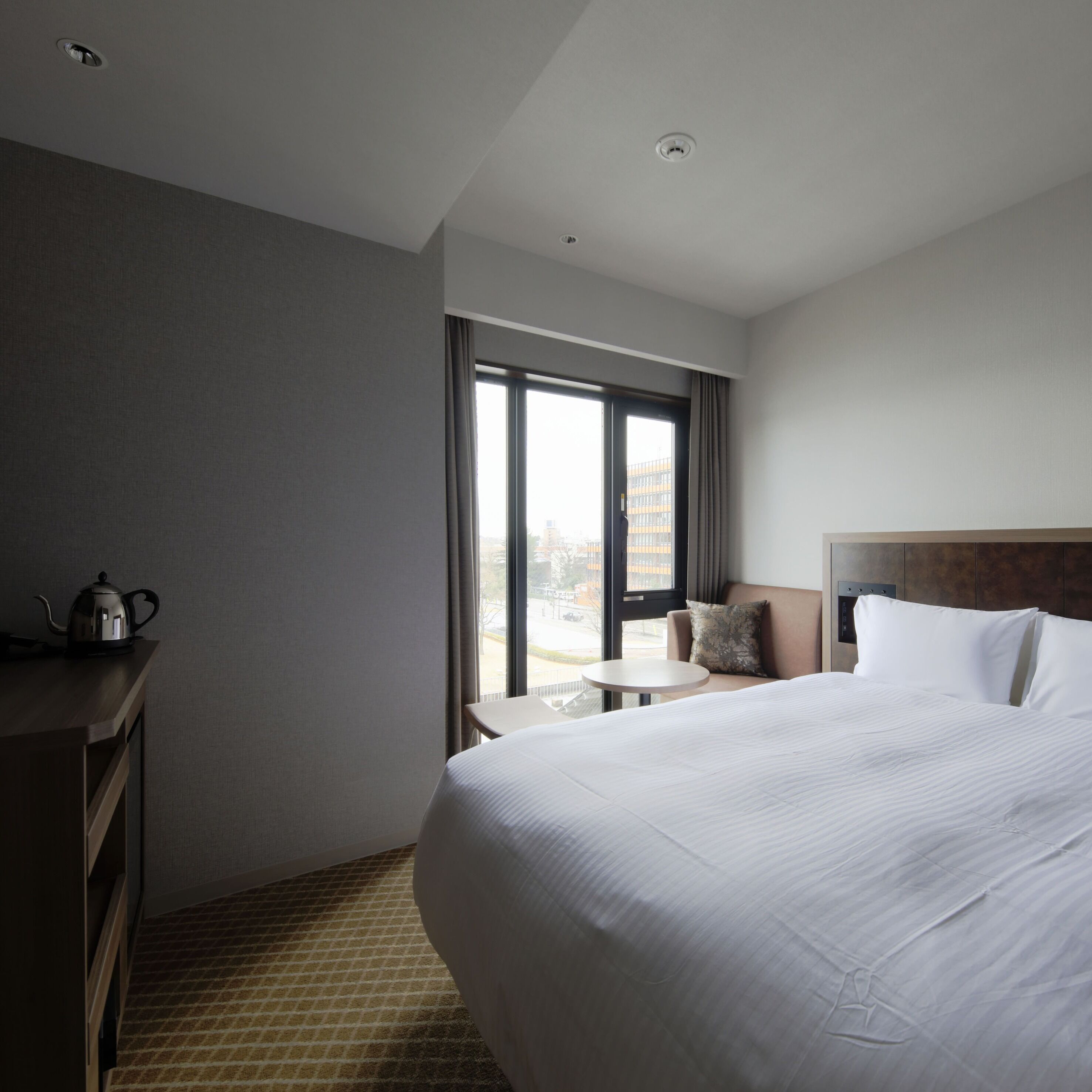 A hotel room with a large bed, a small table, a chair, a desk, and a window with curtains.
