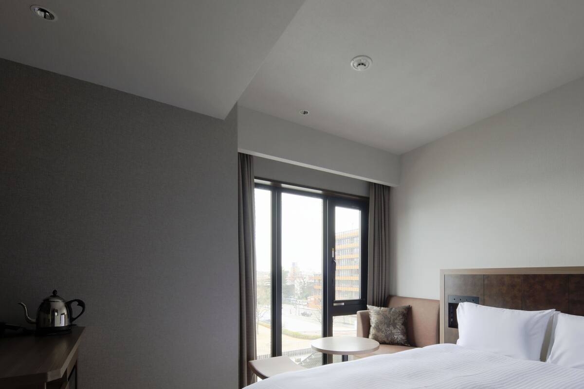 A hotel room with a large bed, a small table, a chair, a desk, and a window with curtains.