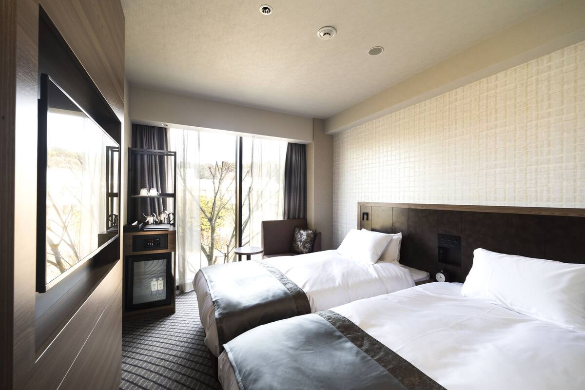 A hotel room with two beds, a desk, a minibar, and a large window with curtains.